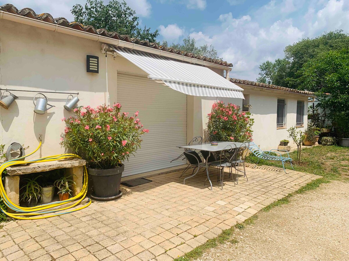 A patio area is featured outside the entrance, with a light-colored stone pathway and a white awning providing shade. A table with chairs is set for dining, surrounded by pots of blooming flowers and greenery, enhancing the outdoor space.