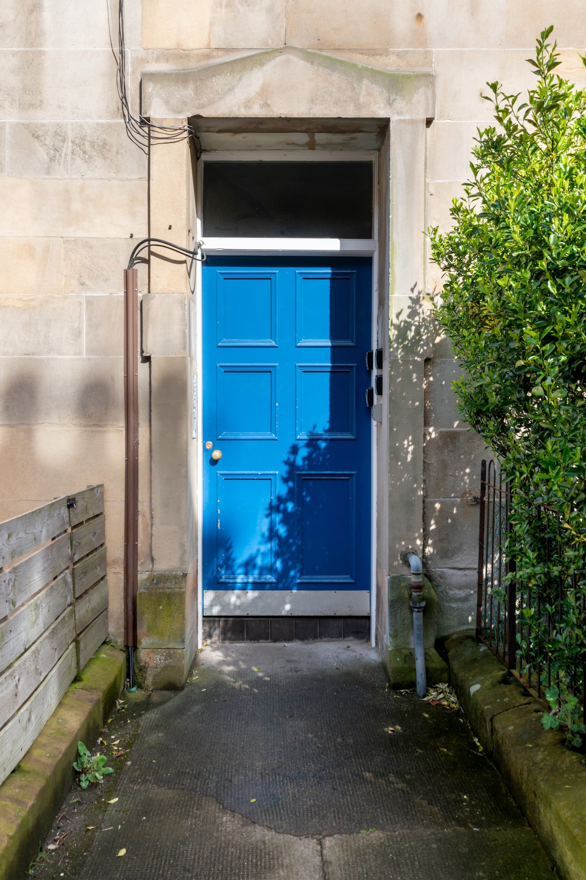 A blue front door is framed by light stone walls, leading to the entrance. Lush greenery is visible on both sides, with a pathway lined by a low wooden fence and a metal railing, creating an inviting approach to the apartment.