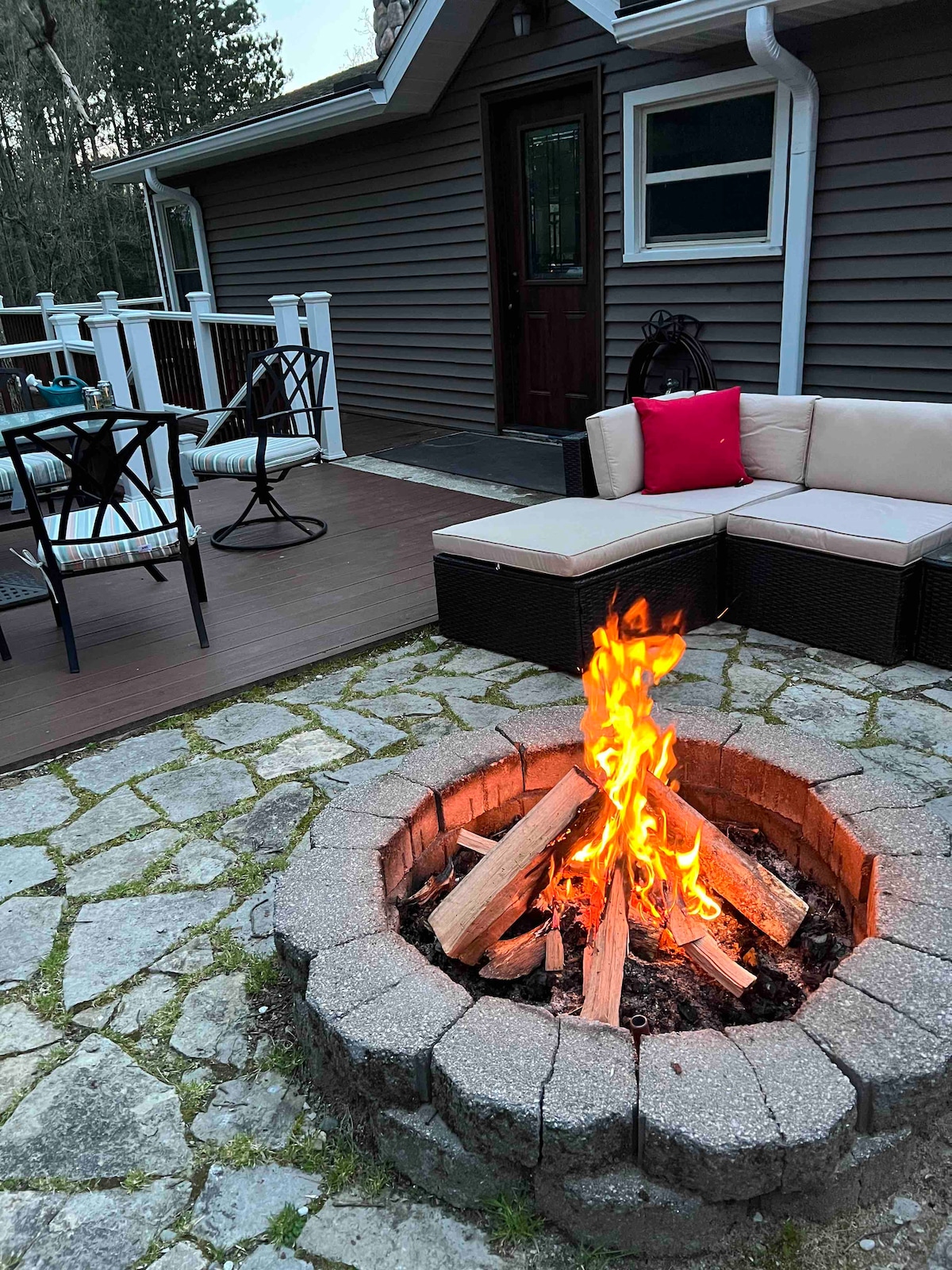 A circular stone firepit filled with arranged firewood radiates warmth. Nearby, a cozy seating area with wicker furniture offers comfortable seating, complemented by decorative pillows. The deck is lined with a wooden railing, and the exterior of the home features a welcoming entryway.