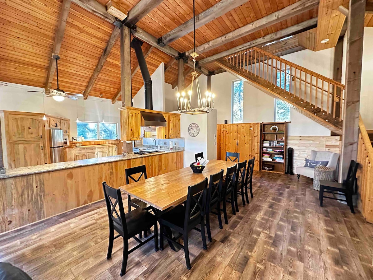 A spacious dining area is highlighted by a long wooden table surrounded by black chairs. The open kitchen features wood cabinetry and granite countertops. Large windows allow natural light to fill the room, and wooden beams accentuate the vaulted ceiling.
