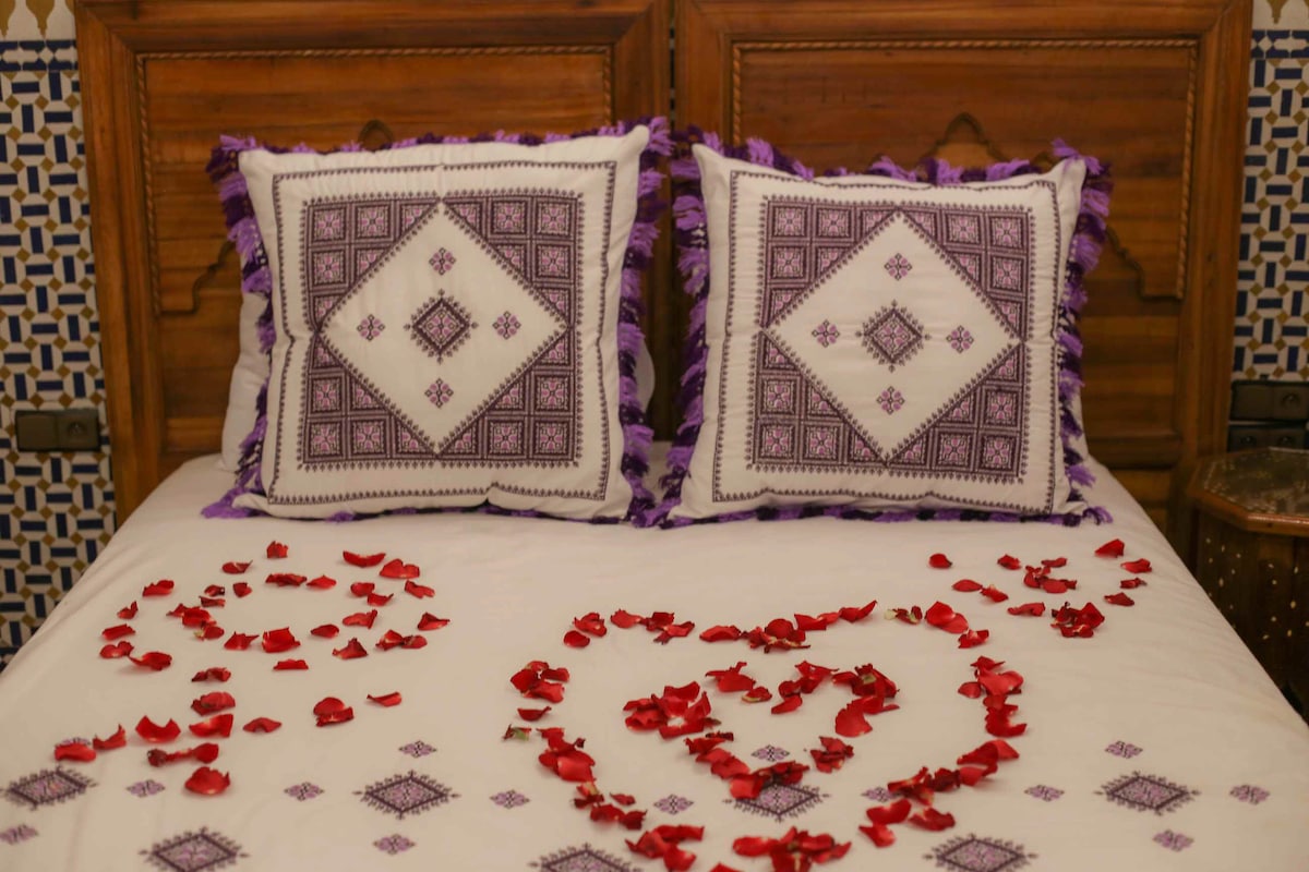 A spacious bed is adorned with a white coverlet featuring decorative patterns. Two purple-fringed pillows are positioned at the head of the bed. Delicate red rose petals are arranged artistically in heart shapes on the bedspread, creating a romantic visual element.