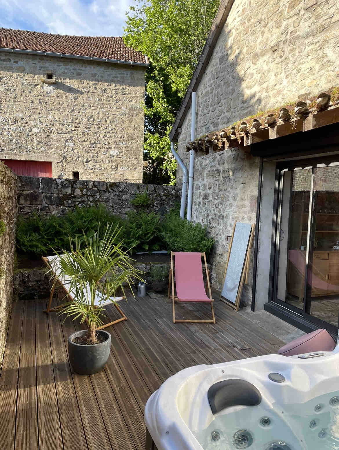 A private outdoor terrace features two lounge chairs placed beside a small potted palm. A spa is positioned nearby, surrounded by a wooden deck, with green foliage visible in the background and stone walls adding texture to the setting.