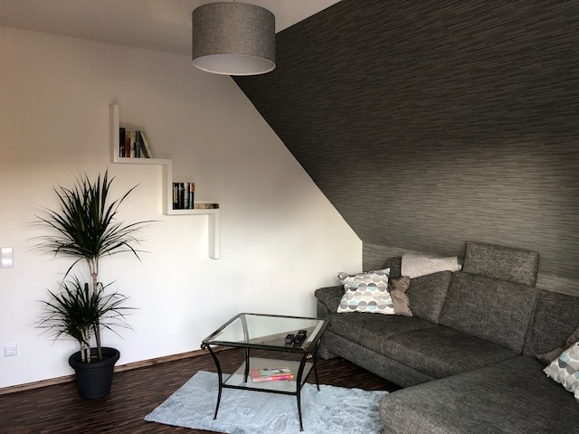 A cozy corner of the living room features a gray sectional sofa arranged around a glass coffee table. A potted plant adds a touch of greenery, while minimalist shelves display books against a light gray wall. Soft lighting is provided by a ceiling fixture.