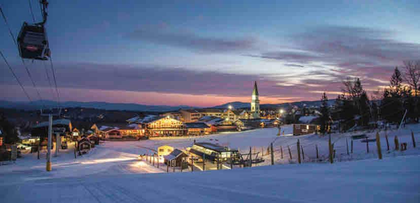 A scenic view captures the Stratton ski area at dusk, showcasing softly illuminated buildings against a colorful twilight sky. The ski lift glides above the snow-covered landscape, leading towards distant mountains. The peaceful scene conveys a tranquil atmosphere for evening activities.