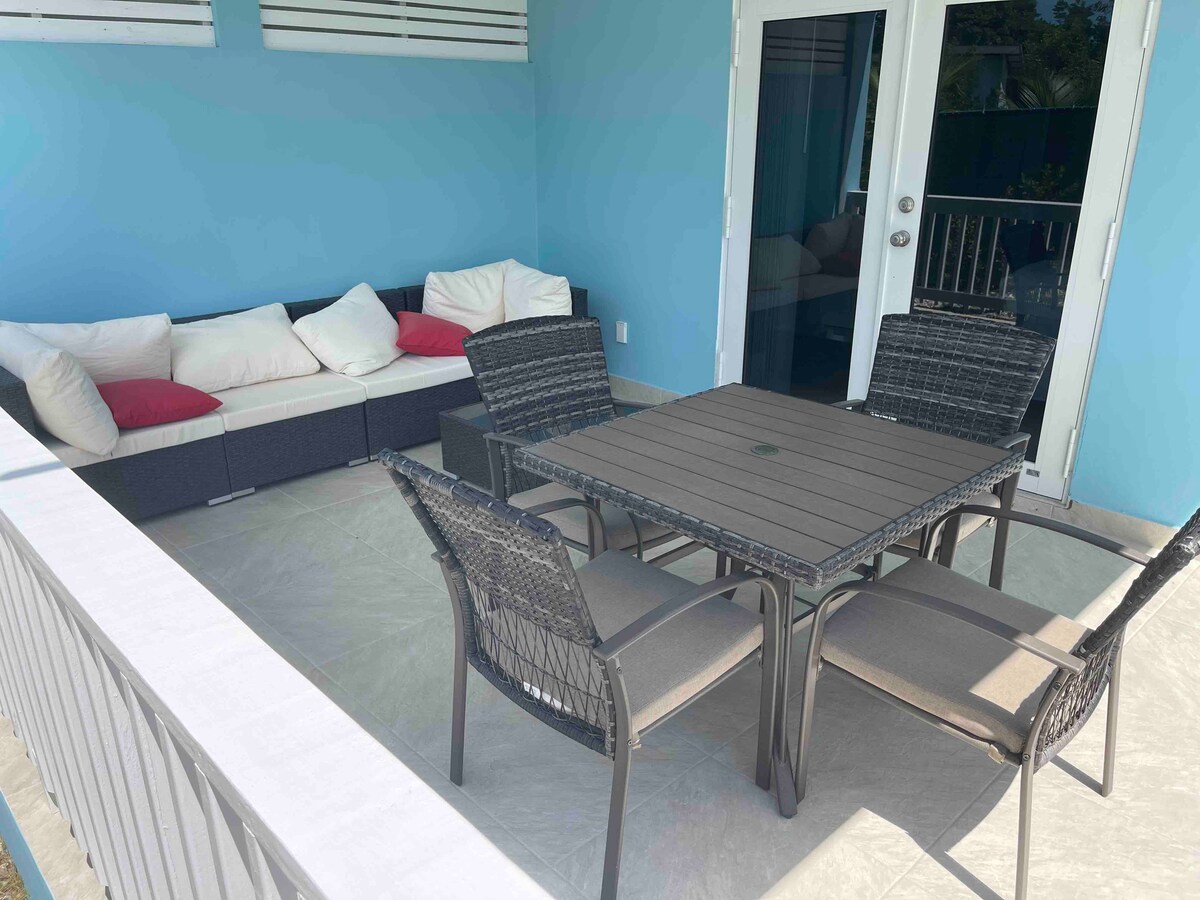 A back patio area features a four-seat dining table accompanied by padded chairs. A sectional sofa with soft cushions is positioned nearby. The space is enclosed with a light blue wall and has large glass doors that allow access to the interior.