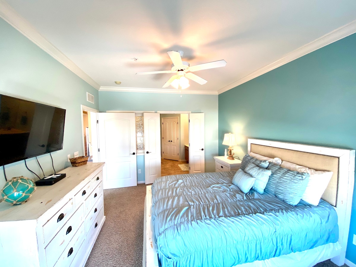 The bright bedroom features light blue walls complemented by a spacious bed dressed in soft bedding. A television is mounted on the opposite wall, and natural light enters through multiple doorways leading to an en-suite bathroom and closet area.