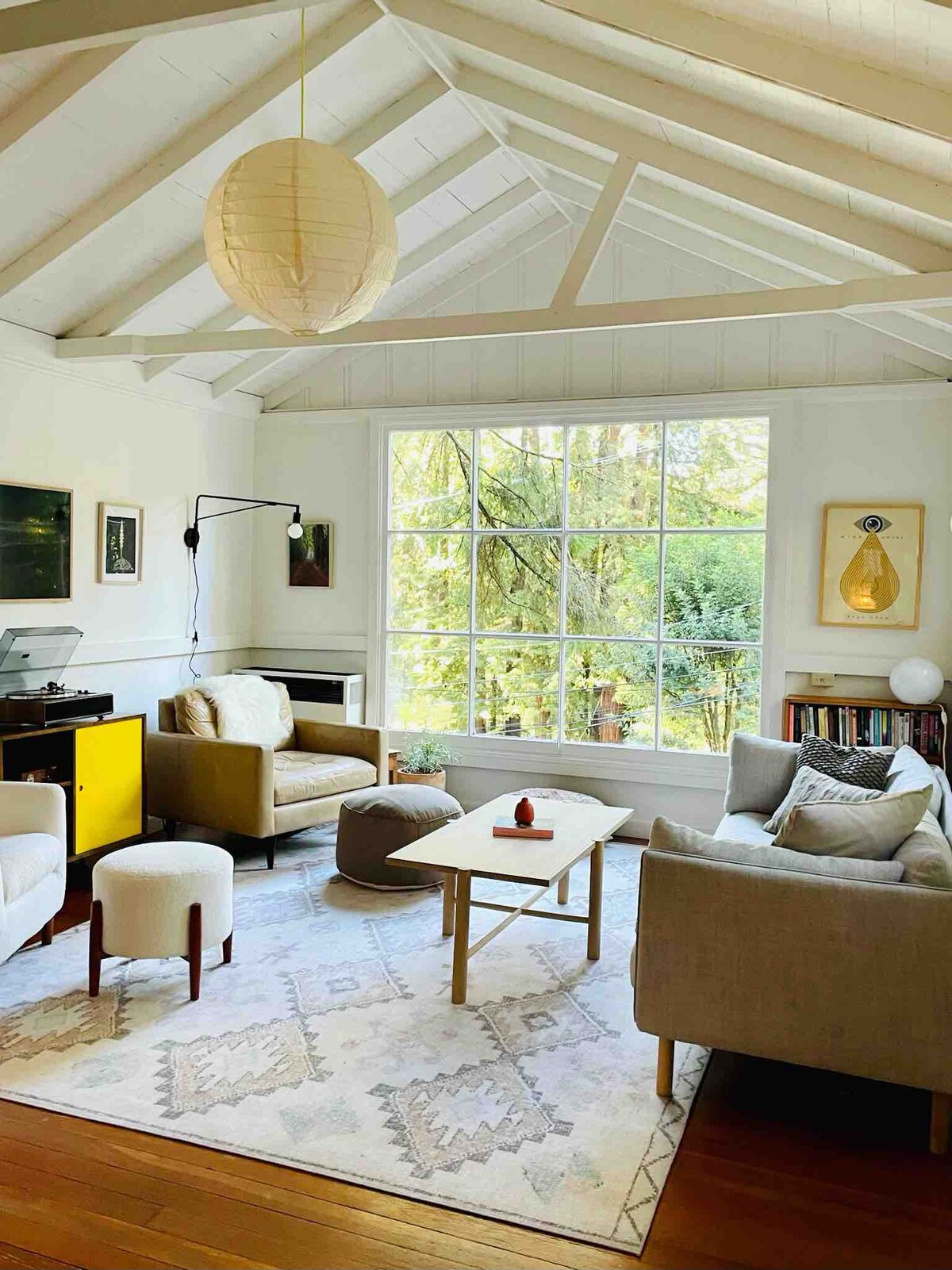 A light-filled family room features vaulted ceilings and large windows overlooking trees. Comfortable seating includes a gray sofa and two armchairs, complemented by a contemporary coffee table. A neutral rug anchors the space, while a stylish pendant light hangs above, adding warmth to the cozy atmosphere.
