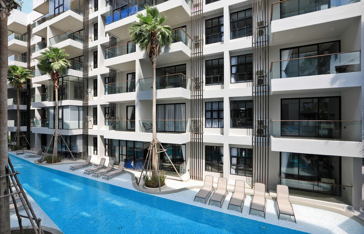 The modern condominium complex features a swimming pool surrounded by palm trees and loungers, providing a relaxing outdoor space. Floor-to-ceiling windows illuminate the apartments, offering views of the pool and lush greenery. The clean lines and neutral tones enhance the contemporary aesthetic.