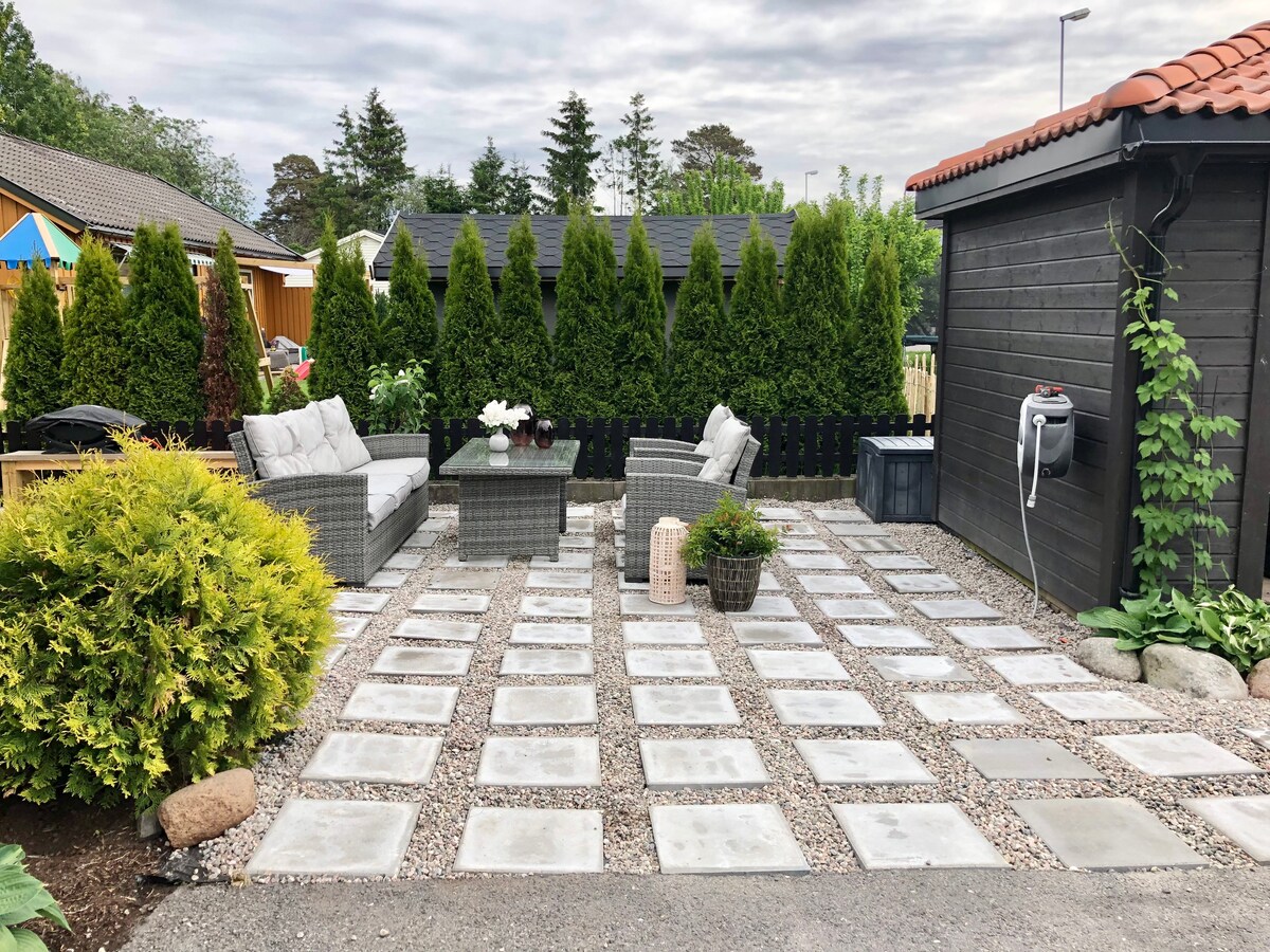 An outdoor area features a stone pathway and is surrounded by tall green hedges. Comfortable seating is provided with two armchairs placed beside a small table, creating a welcoming space. Decorative plants and a lantern add subtle charm to the serene setting.