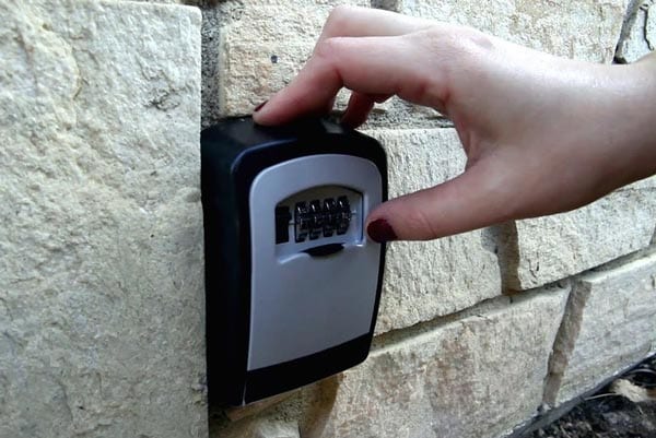 A security lockbox is mounted on a textured stone wall, featuring a combination keypad. A hand is shown adjusting the combination, indicating the accessible entry method to the accommodation. The surrounding area is softly illuminated, enhancing visibility of the lockbox.