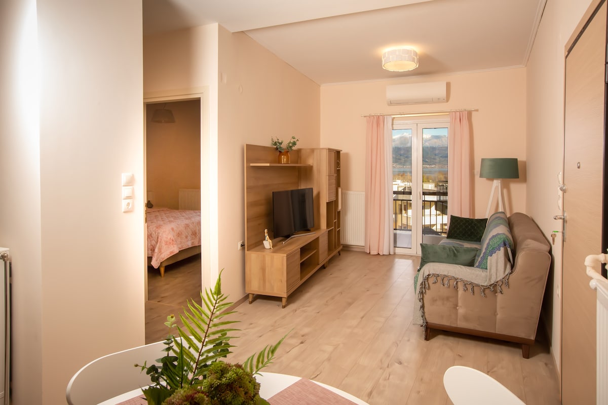 A living area features a comfortable sofa accompanied by a wooden entertainment unit with a television. Light streams through a balcony door, illuminating the space. A small dining table with a plant centerpiece is visible, enhancing the inviting atmosphere of the room.