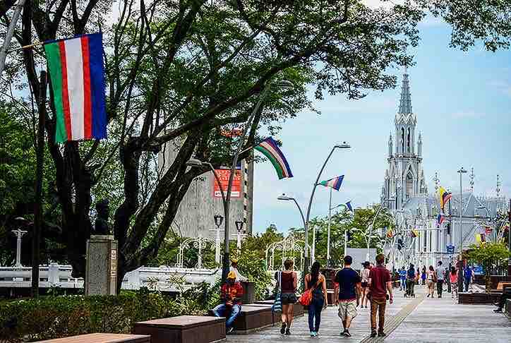 A lively street scene in a vibrant city center is depicted, featuring pedestrians strolling under tall trees. Colorful flags line the pathway, while a prominent church with a tall spire is visible in the background, enhancing the cultural ambiance of the area.