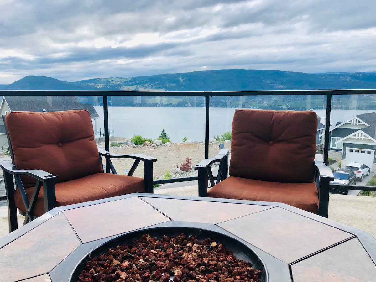Two comfortable chairs are positioned around a firepit on a spacious deck, overlooking Okanagan Lake. The glass railing provides an unobstructed view of the lake and surrounding landscape, inviting relaxation and enjoyment of the scenic vistas.