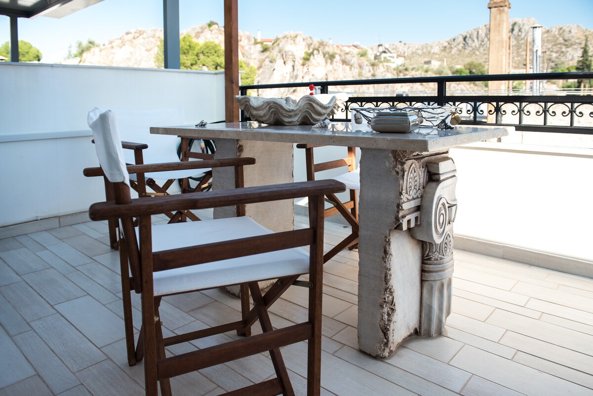 A spacious balcony features a modern table made of stone, accompanied by four wooden chairs with light-colored cushions. The railing offers a scenic view, while rocky hills can be seen in the background, providing a nice contrast to the decor.