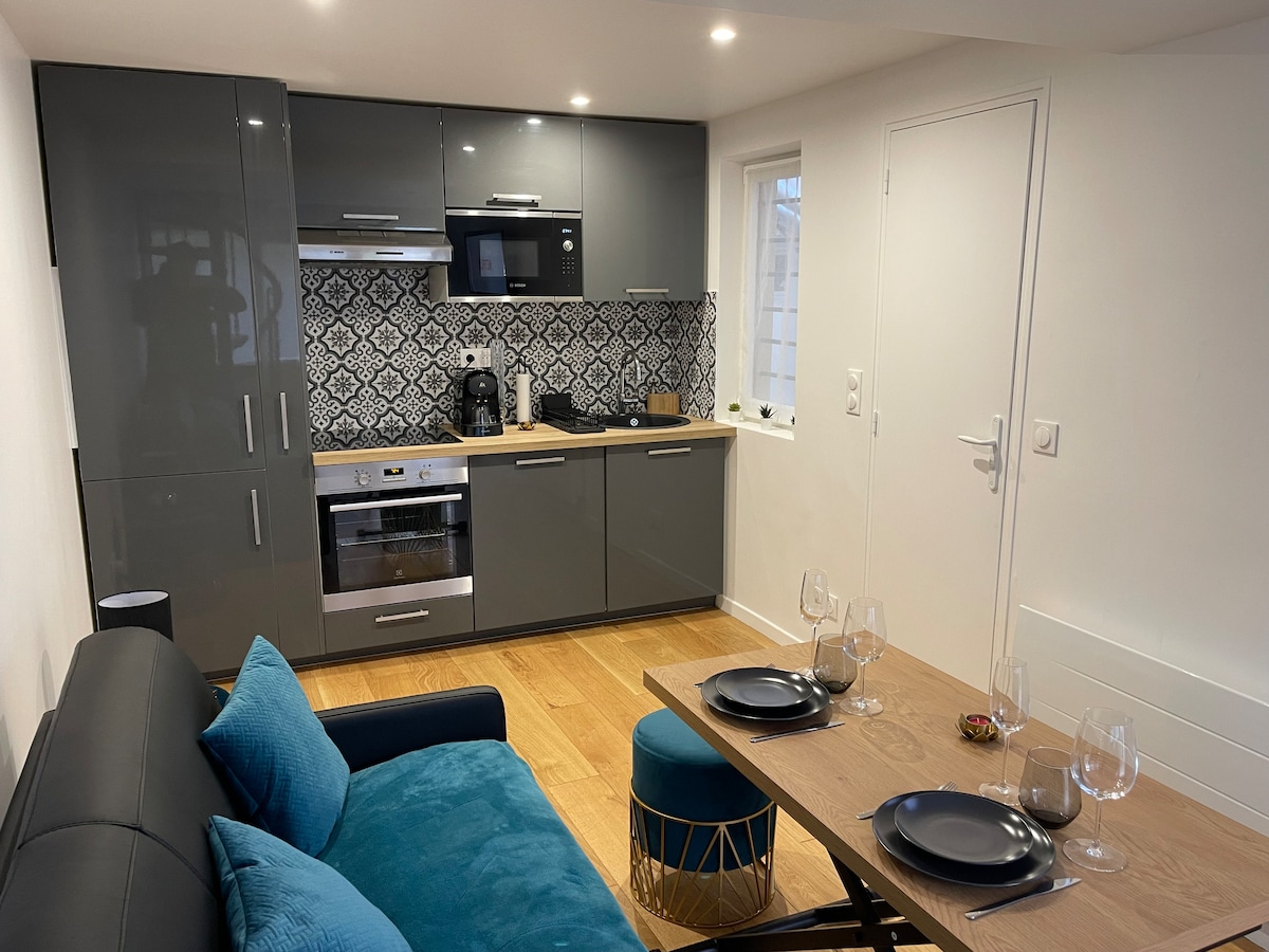 A modern kitchen is presented with sleek gray cabinets and a decorative backsplash. Kitchen appliances, including an oven and coffee maker, are clearly visible. A dining table with two plates and wine glasses is positioned next to a cozy seated area, featuring a colorful blue couch.