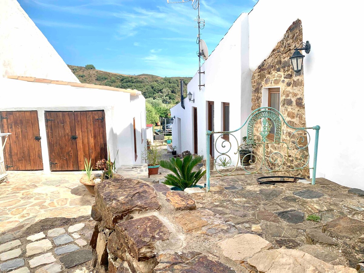 A stone pathway leads through the rustic area, flanked by whitewashed buildings and wooden garage doors. Vibrant greenery, including potted plants, is visible, while a decorative wrought iron gate adds charm to the entrance. Surrounding hills can be glimpsed in the background under a clear sky.