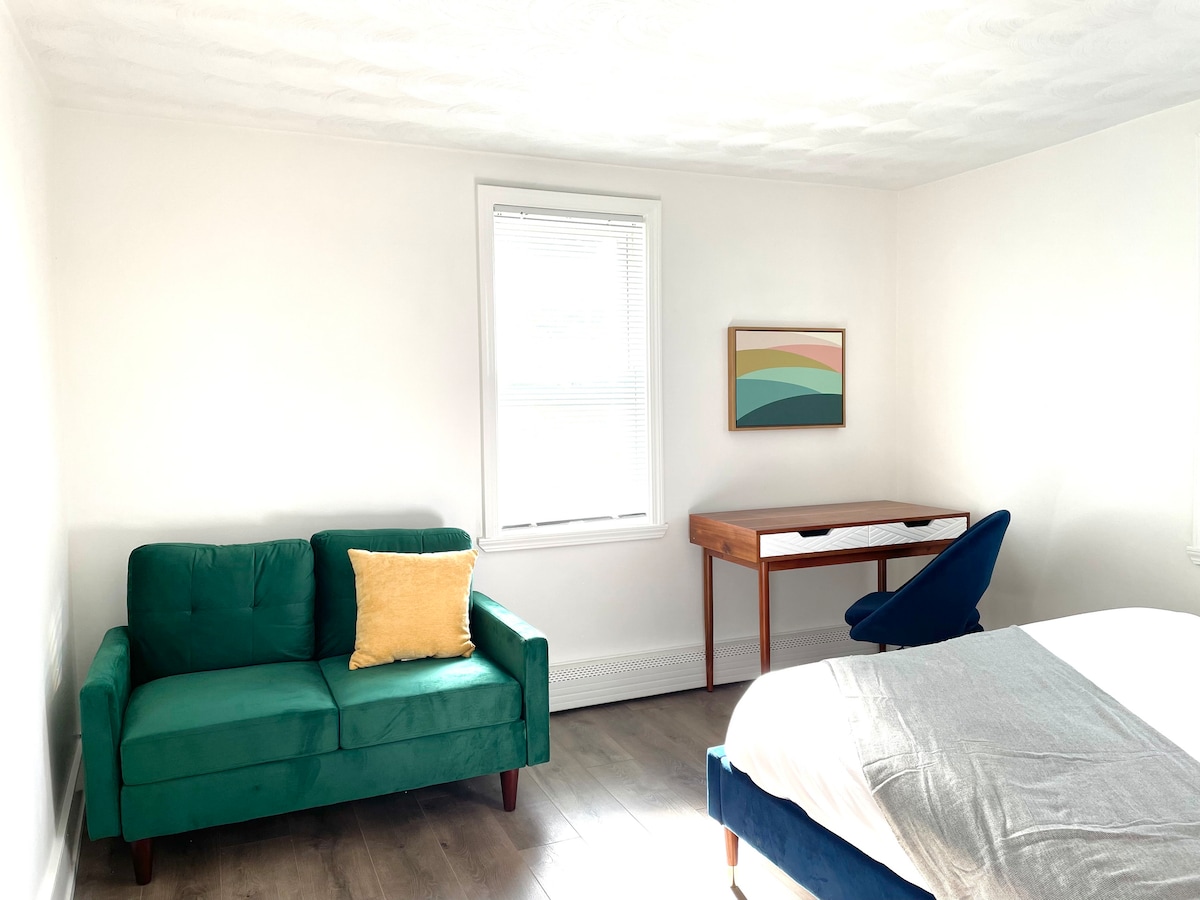 A bright room features a spacious bed covered with light bedding. A green sofa with a yellow cushion is positioned beside a wooden desk and chair. Natural light enters through a window, and a colorful wall art piece enhances the decor.