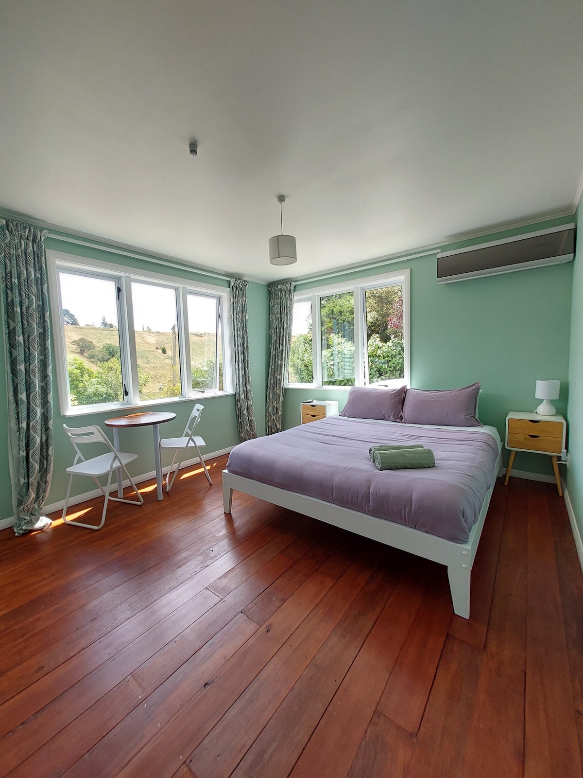 A bright bedroom is presented with large windows that offer views of the surrounding nature. A spacious bed with soft linens is positioned alongside two nightstands. A small dining table and two chairs are placed in the corner, providing additional seating for guests.