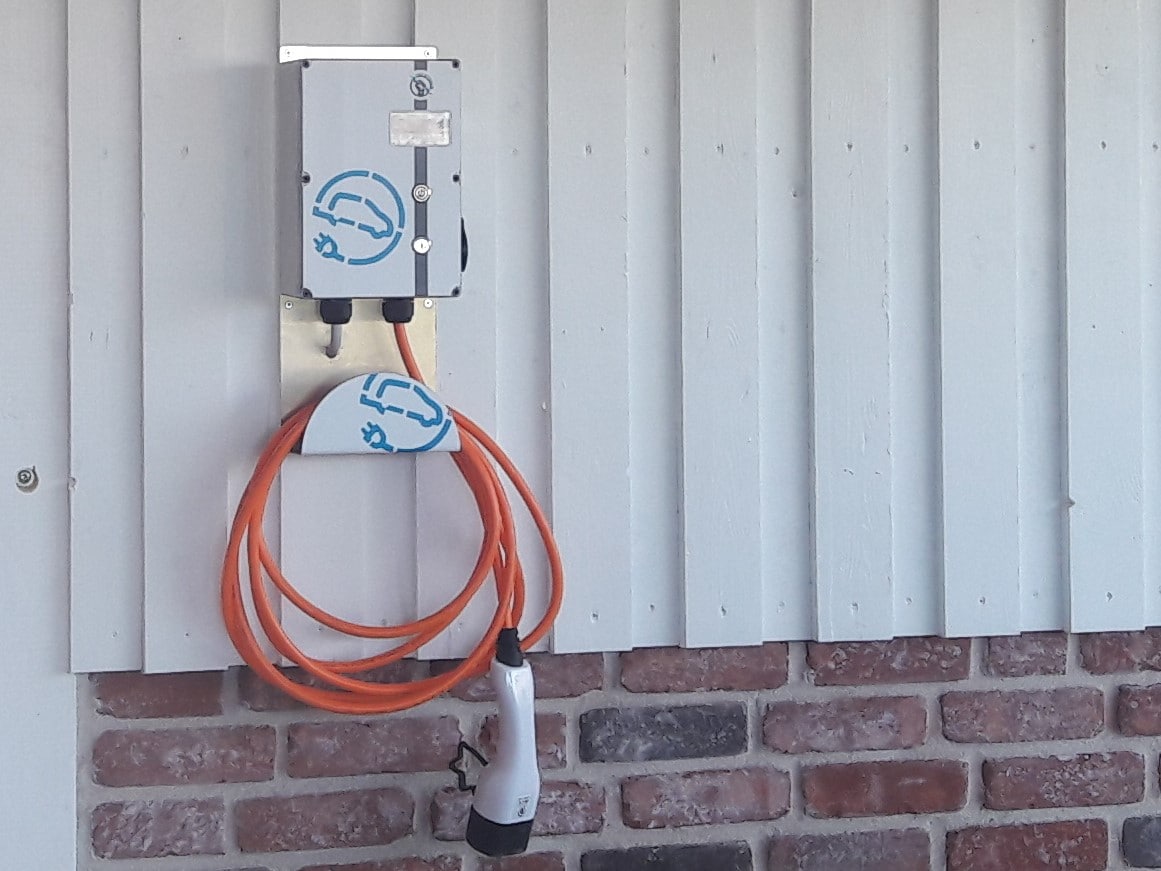 An electric vehicle charging station is mounted on a wooden wall, featuring a white cord neatly wrapped around the unit. The station is positioned beside a section of exposed red brick, providing a functional amenity for guests with electric vehicles.