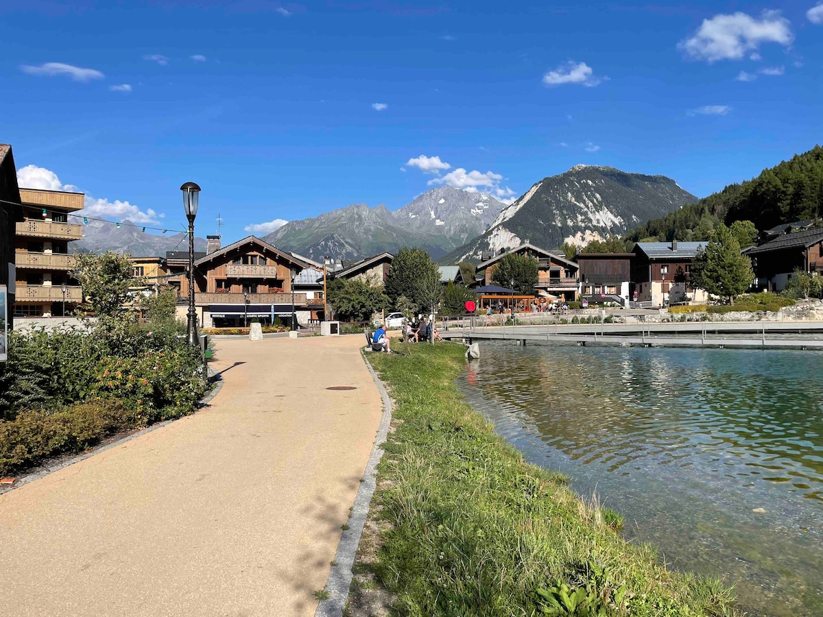 A scenic pathway runs alongside a calm lake, surrounded by residential buildings and lush greenery. Majestic mountains rise in the background under a clear blue sky, creating a serene and inviting atmosphere for leisurely walks.