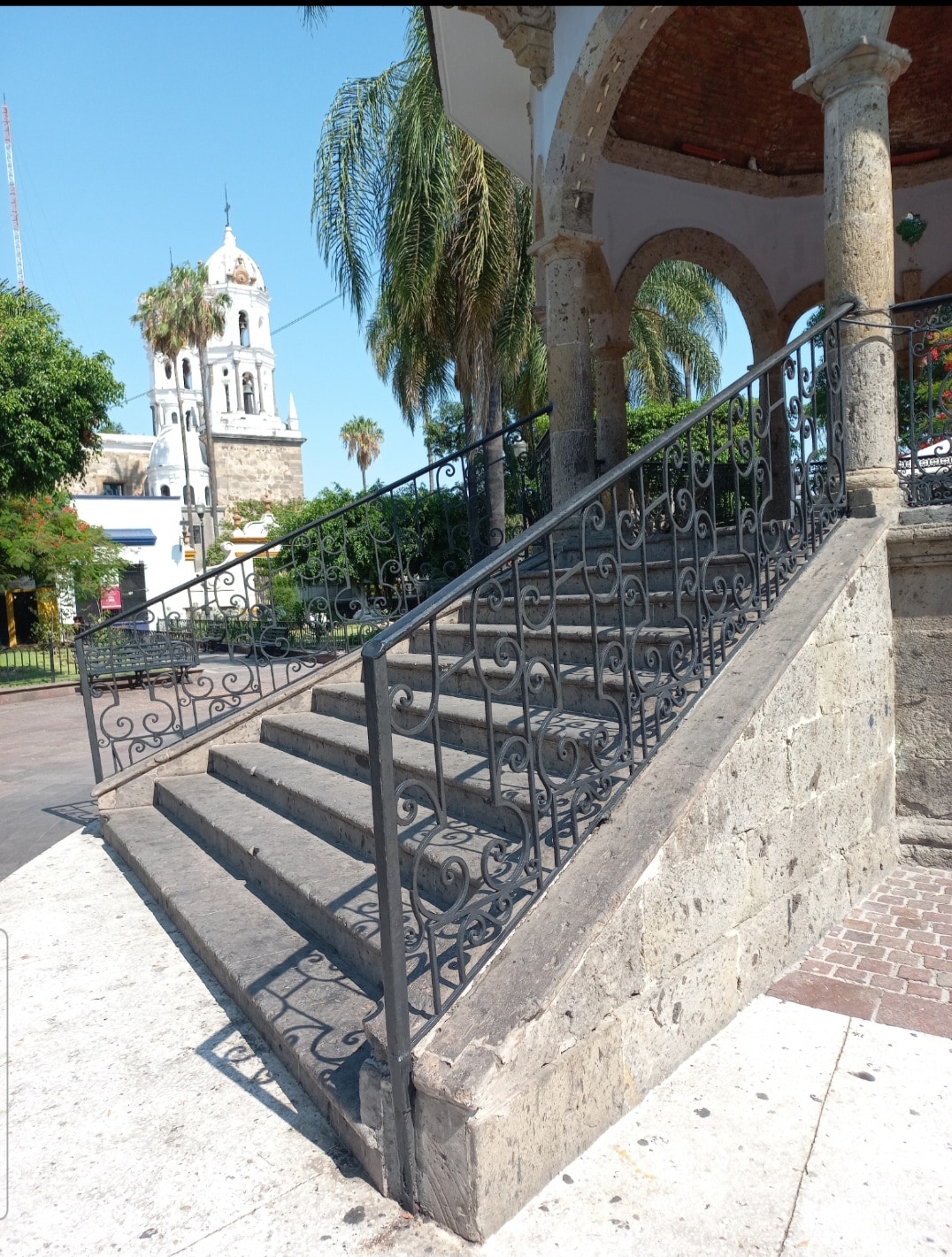 A set of stone stairs with decorative wrought iron railings leads up to a plaza area. Lush palm trees and historical buildings provide a scenic backdrop. In the distance, a tower with a bell and ornamental architecture can be seen, reflecting the charm of the surrounding area.