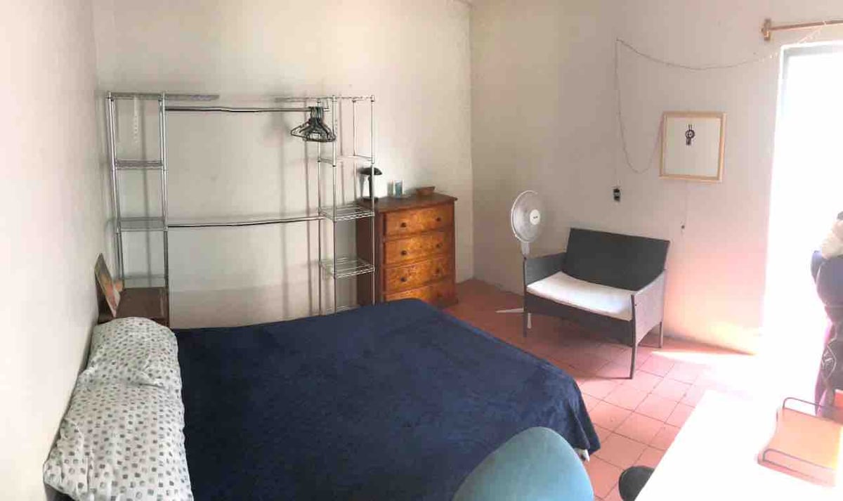 A spacious room features a large bed covered with a navy blue blanket and patterned pillows. Nearby, a wooden dresser is situated along with a fan and a clothing rack. A gray sofa and a simple desk complete the space, with natural light illuminating the area.