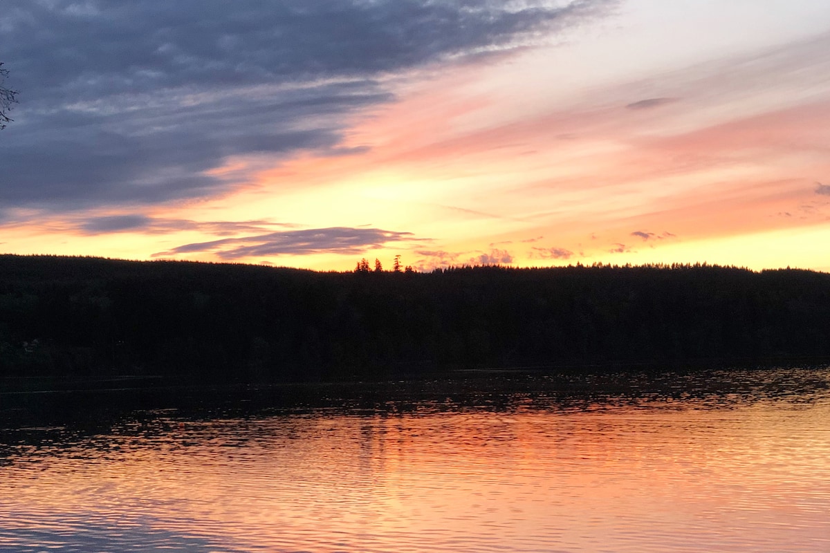 An evening sky transitions from soft hues of orange and pink to deep purples, reflecting on the still water below. Silhouetted trees line the horizon, creating a serene backdrop against the colorful sky.