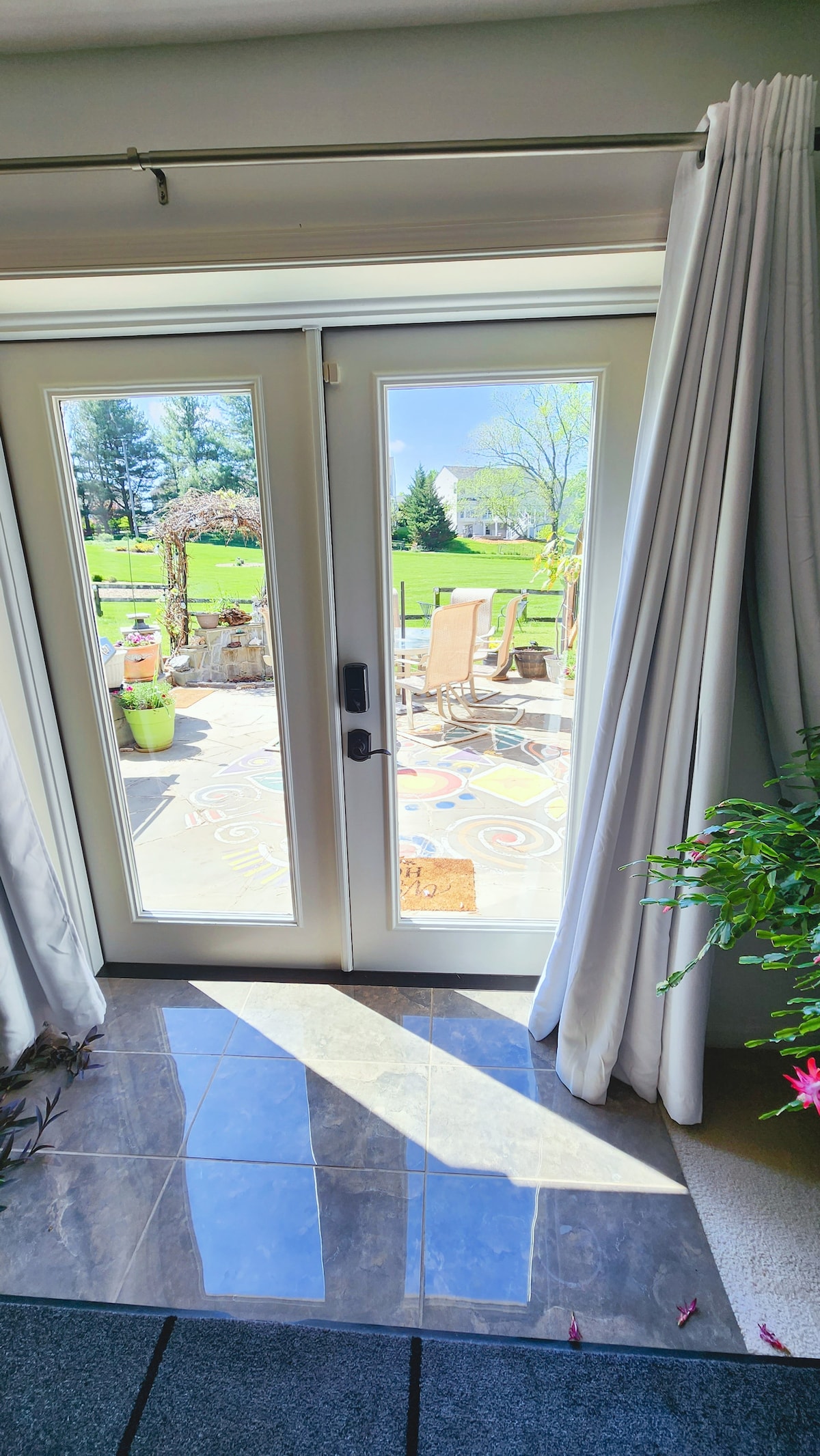 Sliders lead to a patio from the spacious living area, framed by sheer curtains. A well-maintained outdoor space is visible, featuring chairs and a table on a colorful patterned surface. Sunlight reflects on the tiled floor near the entrance.
