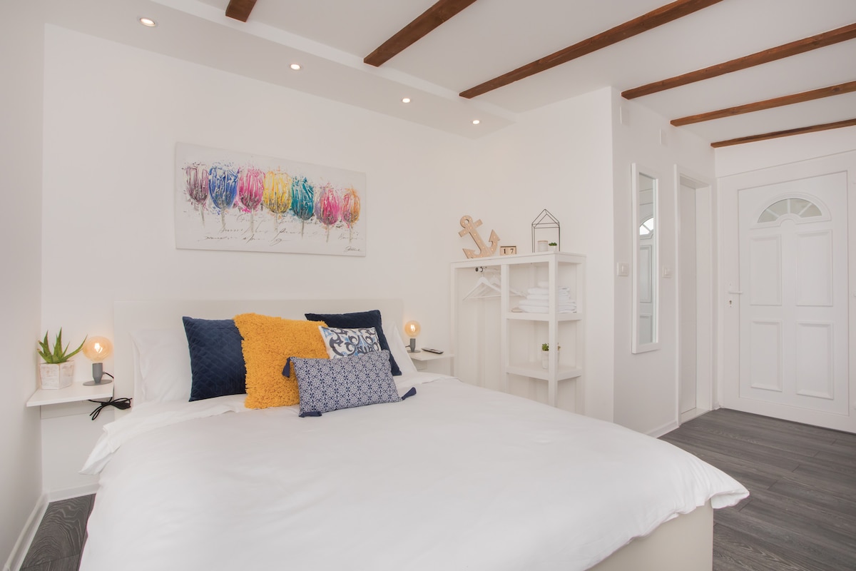 A bright sleeping area features a spacious bed dressed in white linens, complemented by colorful decorative pillows. A vibrant art piece hangs above the bed, while natural light filters in through a nearby door with a frosted glass panel. Simple furnishings enhance the modern design.