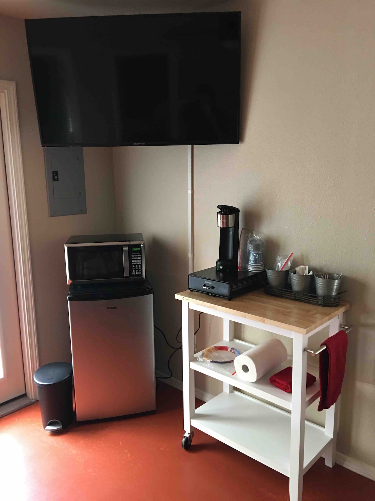 A compact kitchenette area is showcased, featuring a silver microwave and a sleek mini-fridge. A flat-screen TV is mounted on the wall above. A mobile cart holds a coffee maker, utensils in cups, napkins, and paper towels, providing essential amenities for guests.