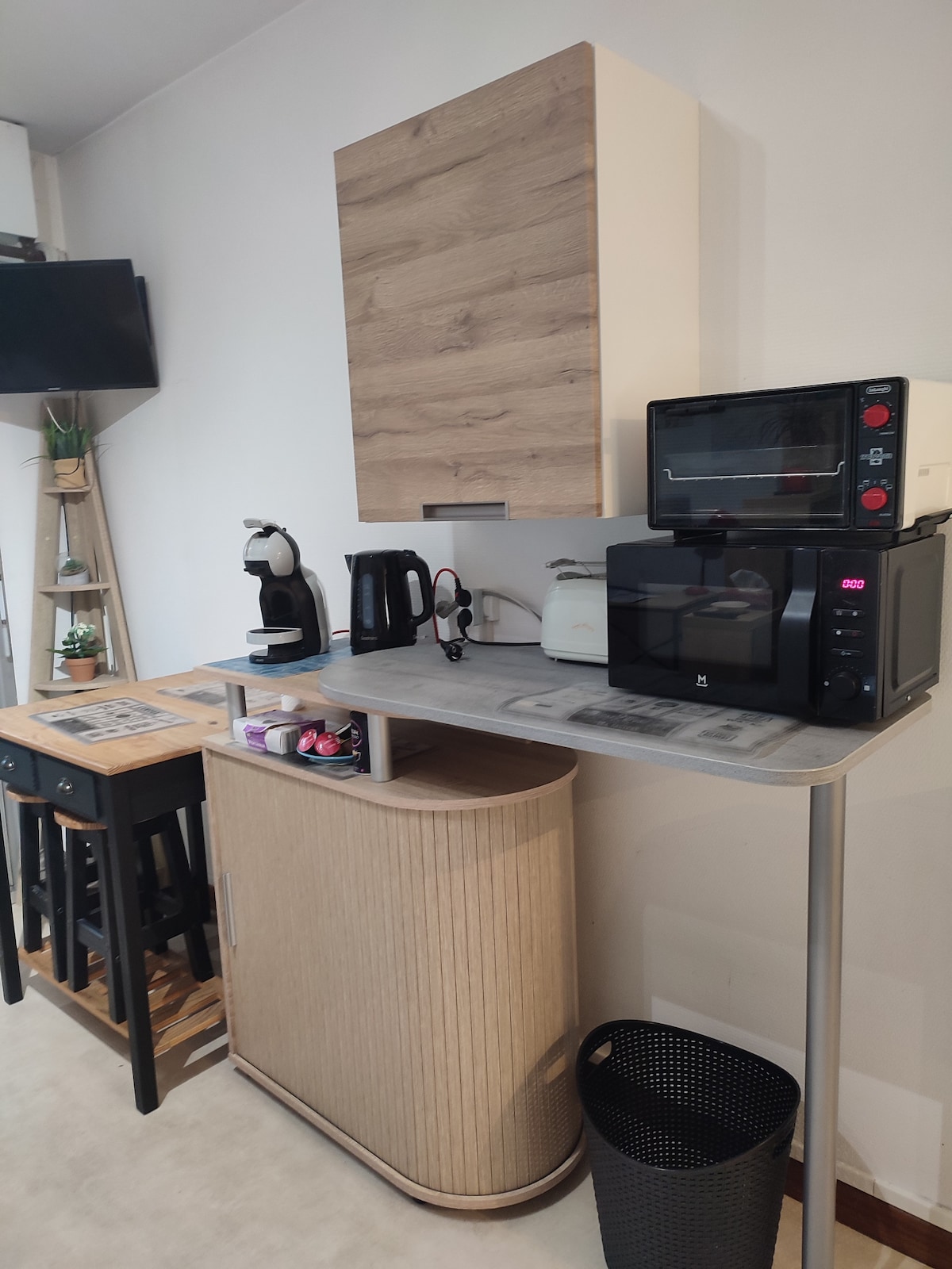 A compact kitchen area features a modern countertop with appliances including a microwave, kettle, and toaster. A sleek cabinet houses additional items, while bar stools are placed nearby. A plant and a wastebasket enhance the overall functionality of the space.
