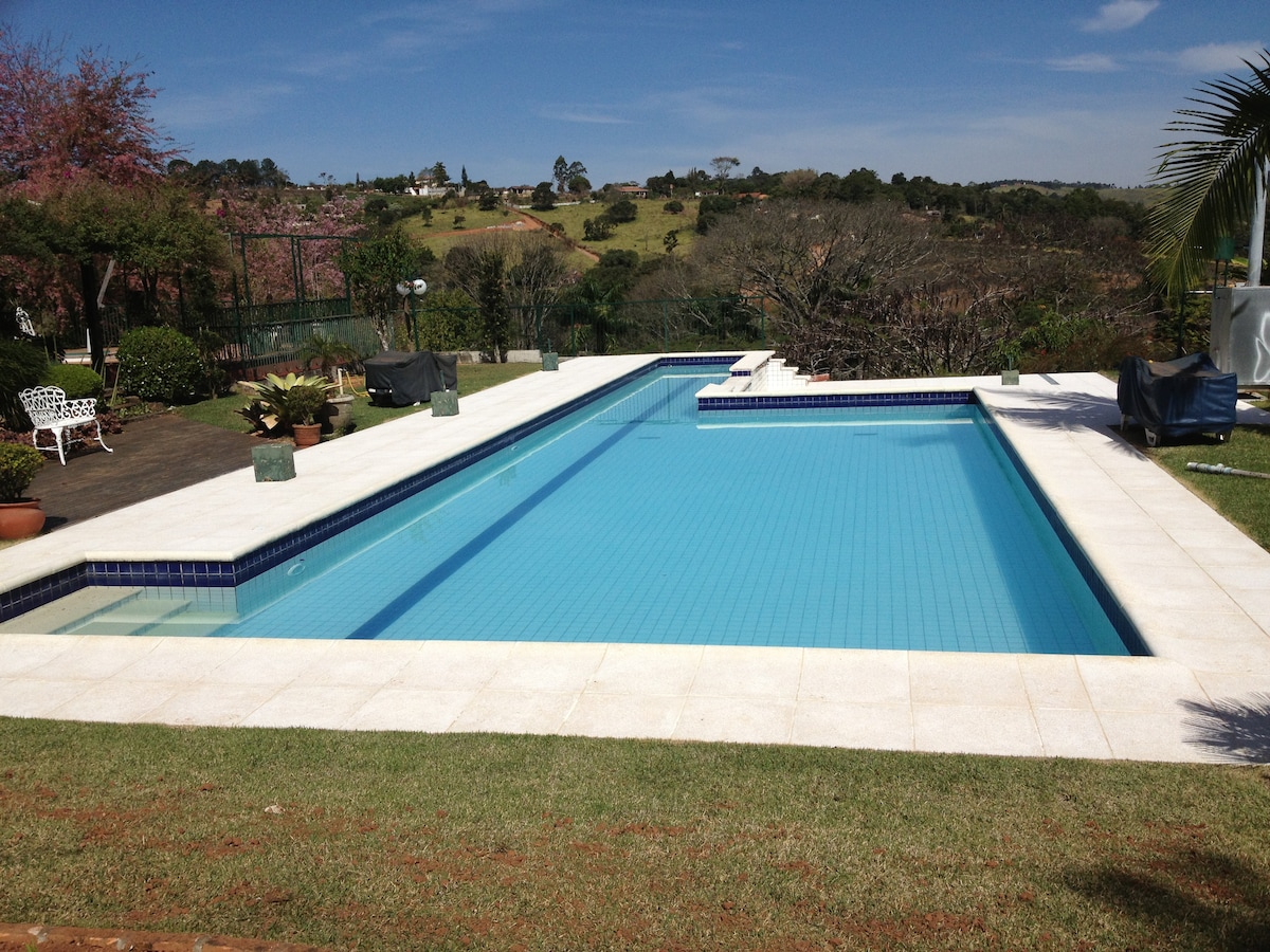A spacious swimming pool with a section for children is surrounded by a sunlit patio. Lush greenery and distant hills are visible in the background, while lounge chairs are positioned for relaxation along the poolside. A mix of potted plants enhances the outdoor ambiance.