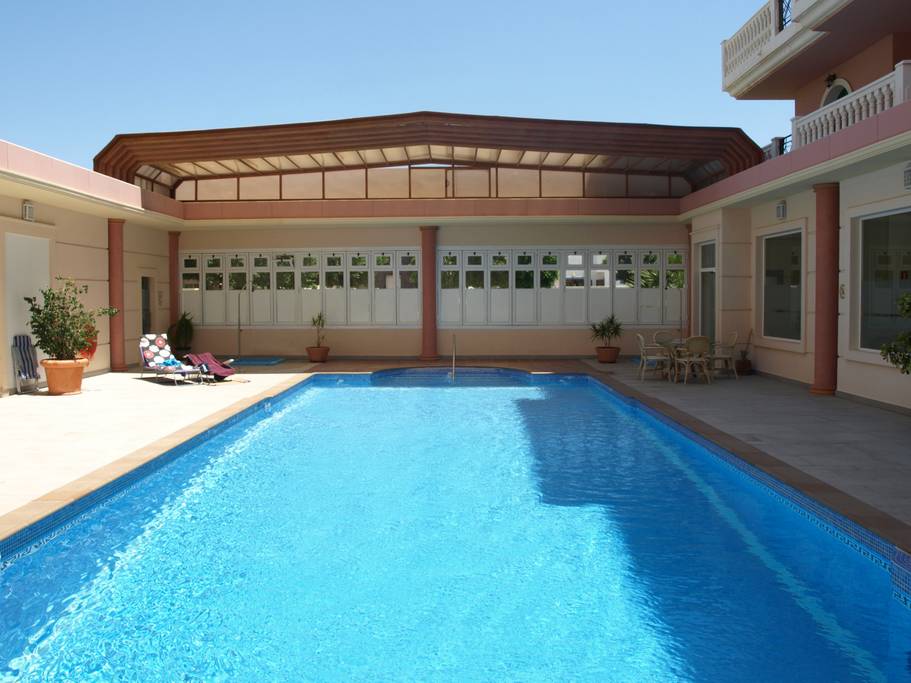 A heated swimming pool is surrounded by a spacious patio, featuring sun loungers and potted plants. The area is bathed in natural light, with large windows providing a clear view. The pool surface reflects the blue sky, creating a serene atmosphere.