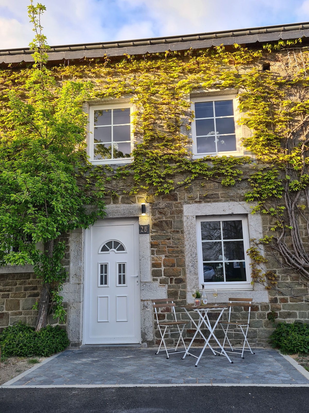 The façade of a stone house is adorned with climbing greenery. Two white chairs and a small round table are positioned on a paved area in front, inviting relaxation. Two windows above the entrance provide natural light, complemented by a white door.