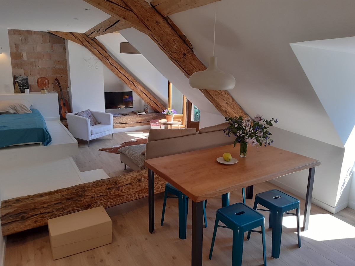 An inviting loft space features exposed wooden beams and a rustic stone wall. A light-colored dining table with blue stools is positioned nearby, while a cozy seating area includes a soft armchair and low table with a flower arrangement. Natural light floods the room.