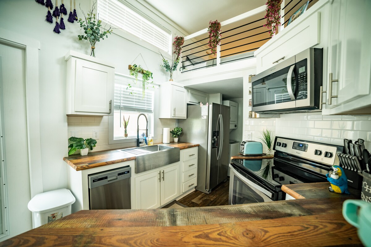 A well-equipped kitchen features modern appliances, including a refrigerator, stove, and microwave. Wooden countertops enhance the space, complemented by light-colored cabinets. Decorative plants and flowers are placed on shelves, and the double sink faces a window, allowing natural light to brighten the area.