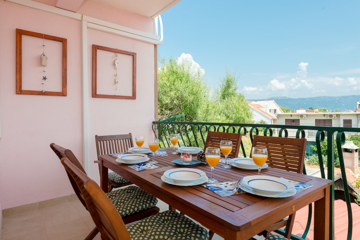 An outdoor dining area is set with a wooden table and eight chairs, arranged for a meal with plates and glasses filled with orange juice. The surrounding balcony features light pink walls and framed decorations, offering a view of greenery and distant rooftops under a clear blue sky.