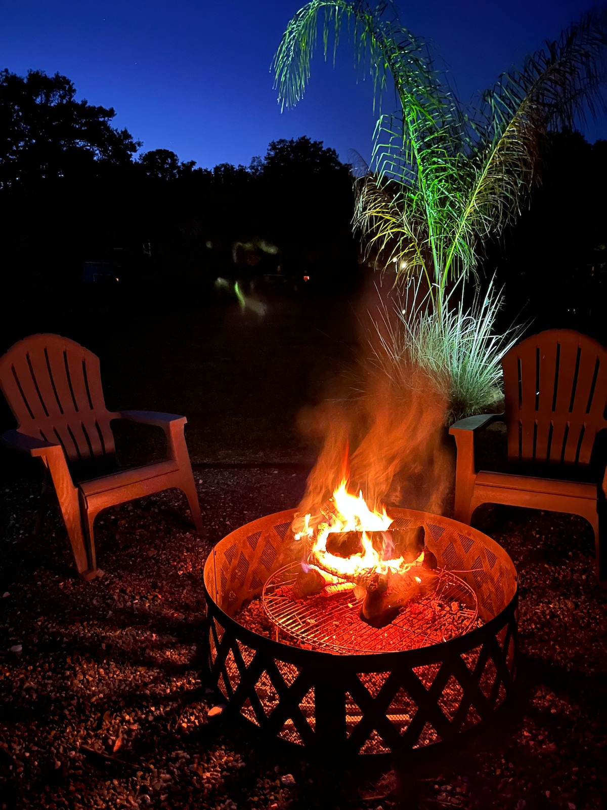 A fire pit is set in a gravel area, surrounded by two wooden Adirondack chairs. Flames dance above the logs, casting a warm glow. In the background, a tall palm tree is illuminated with green light, creating a serene nighttime ambiance.