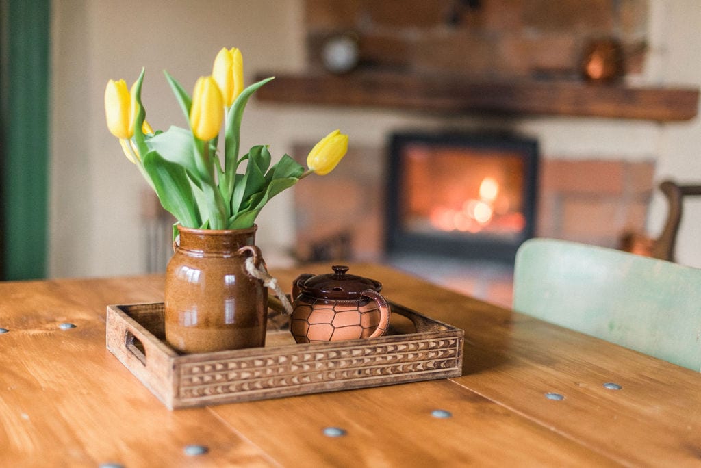 A wooden table is set with a tray containing a decorative teapot and a brown vase filled with fresh yellow tulips. In the background, a cozy fireplace emits a warm glow, enhancing the inviting ambiance of the space.