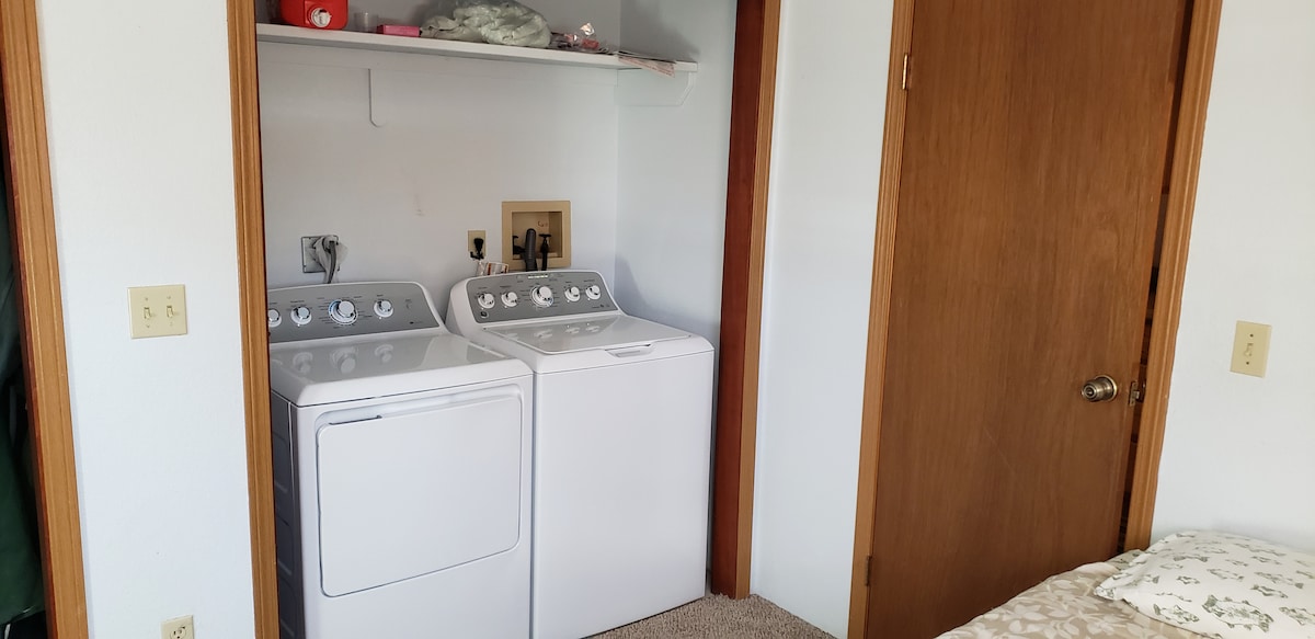 The image displays a separate laundry area featuring a washer and dryer set, positioned adjacent to a bedroom. Above the appliances, shelving is visible, holding various items. The room is finished with cream-colored walls and a carpeted floor.