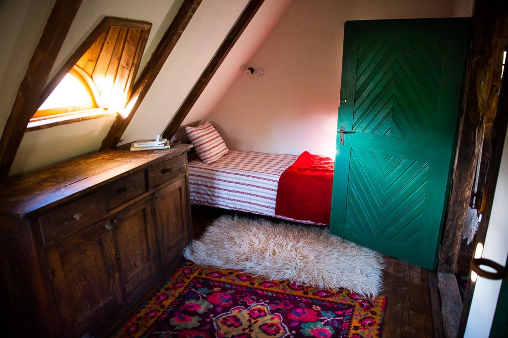 An inviting room features a sloping ceiling, with a single bed dressed in striped linens and a cozy red blanket. A wooden cabinet stands against the wall, while a decorative rug adds warmth to the space. Natural light filters through a small window.