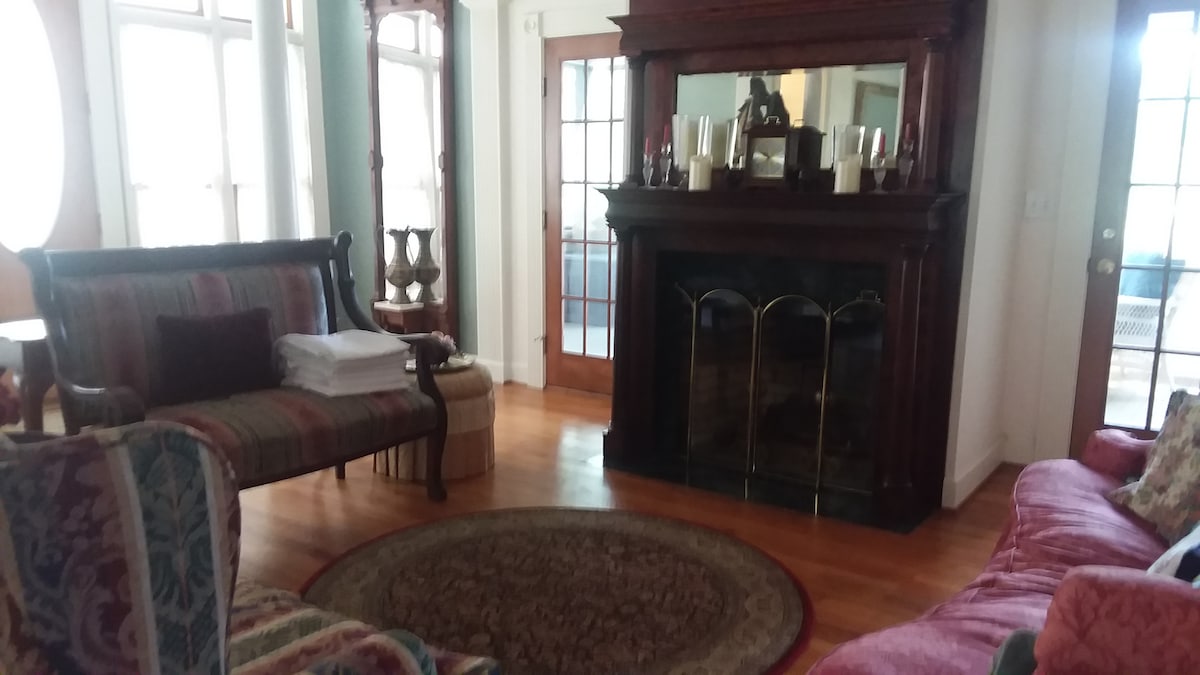A cozy living room showcases a classic wooden fireplace framed by decorative candles. Comfortable seating includes a striped sofa and a plush reddish chair, arranged around a circular area rug. Large windows allow natural light to brighten the space, enhancing its inviting atmosphere.
