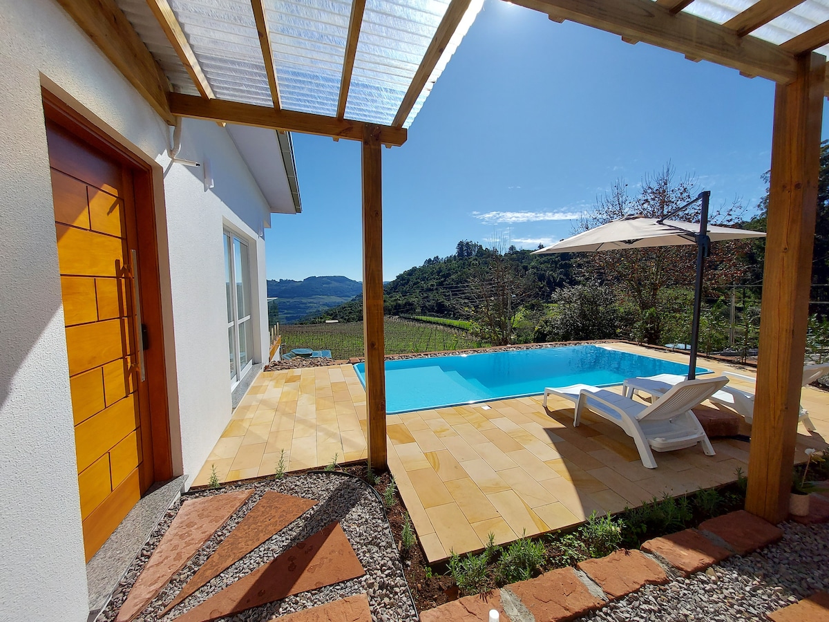 The outdoor area features a private pool with clear blue water, surrounded by a tiled patio. A shaded area with an umbrella and a lounge chair is present. Scenic views of surrounding hills and vineyards can be seen in the background under a bright blue sky.