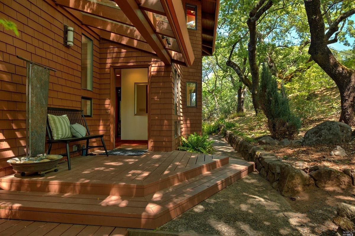 The entrance of the home features a covered porch with wooden decking, framed by natural stone borders and lush greenery. A bench is positioned invitingly, complemented by vibrant outdoor landscaping through the large trees.