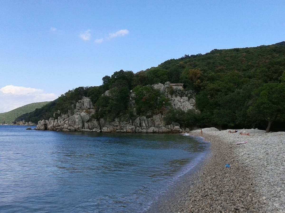 A serene beach is visible, bordered by a pebbly shoreline and azure water. Rocky formations rise from the coastal landscape, accompanied by lush greenery from the hills in the background, creating a natural and tranquil environment.
