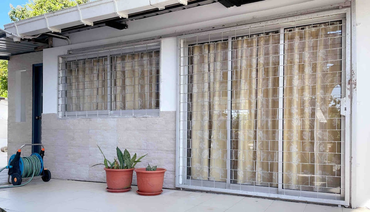 The exterior showcases a small patio area framed by two windows with security grills. Light-colored curtains hang in front of the glass, while two potted plants add a touch of greenery. A garden hose is neatly coiled on the ground, ready for use.