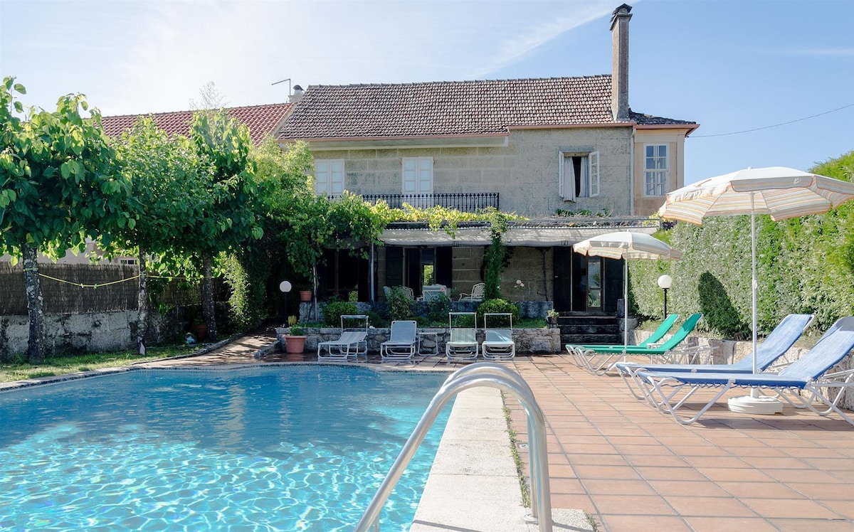 A private outdoor pool area is visible, surrounded by lush greenery and sun loungers. The house features a spacious terrace with multiple seating areas, providing a welcoming space for relaxation. The sun shines brightly, enhancing the serene atmosphere of the garden.