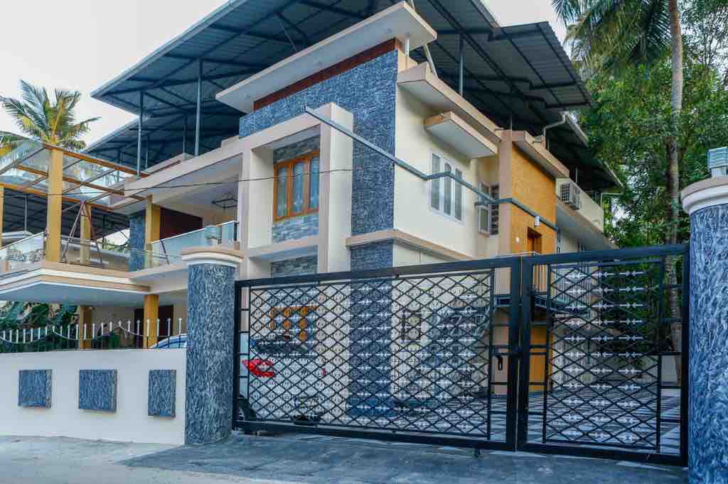 A modern villa is showcased within a gated entrance, highlighting a mix of stone and stucco textures. The structure features multiple levels with balconies, surrounded by palm trees, and a parking area visible nearby.