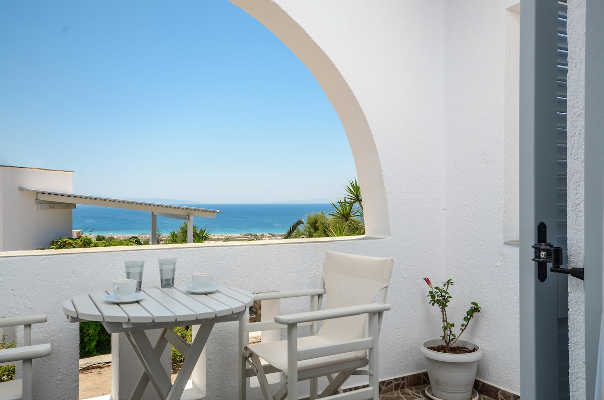 A private shaded balcony is depicted, featuring a small round table with two glasses and chairs. An arched window frames a view of the sea and distant land, while greenery adds a natural touch to the scene.