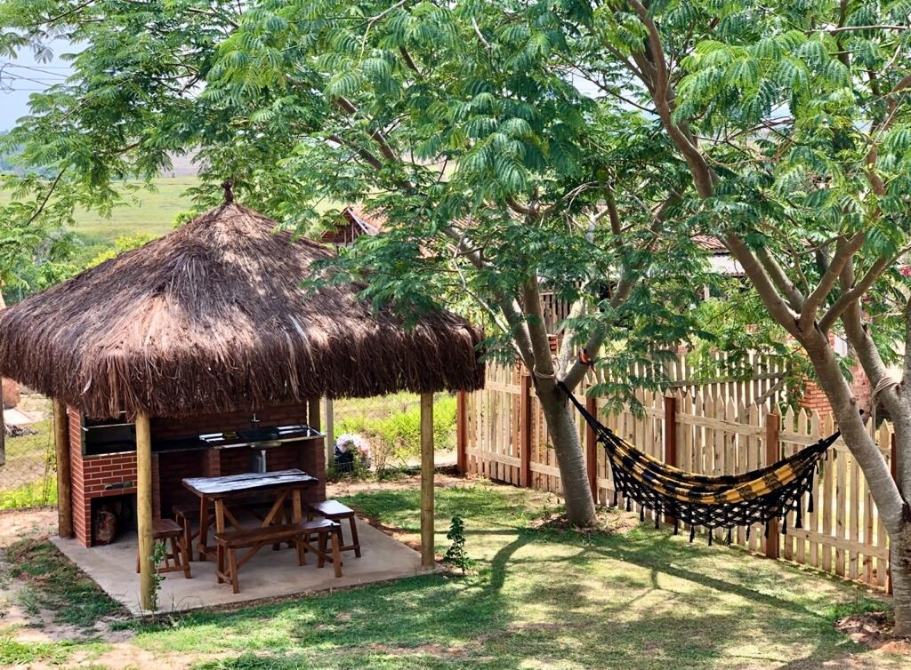 A shaded outdoor space features a thatched-roof gazebo with a wooden dining table and benches. Nearby, a hammock is strung between trees, providing a cozy spot to relax. The lush green lawn is surrounded by a wooden fence, enhancing privacy.